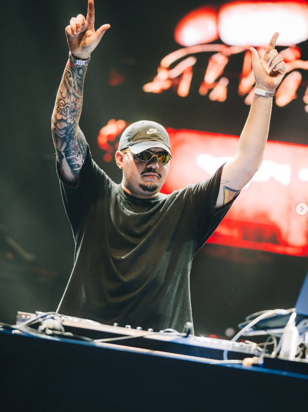 DJ Wallow com os dois braços para o alto diante de um telão vermelho